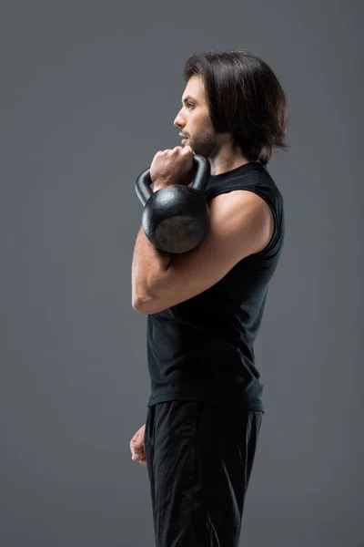 Вид сбоку на мускулистого молодого человека, упражняющегося с гирей, изолированного на сером — стоковое фото