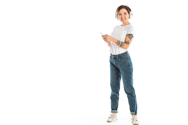 Mujer joven sonriente que usa auriculares, escucha música y usa un teléfono inteligente aislado en blanco - foto de stock