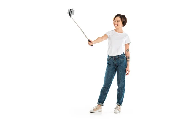 Beautiful young woman taking selfie on smartphone using selfie stick isolated on white — Stock Photo