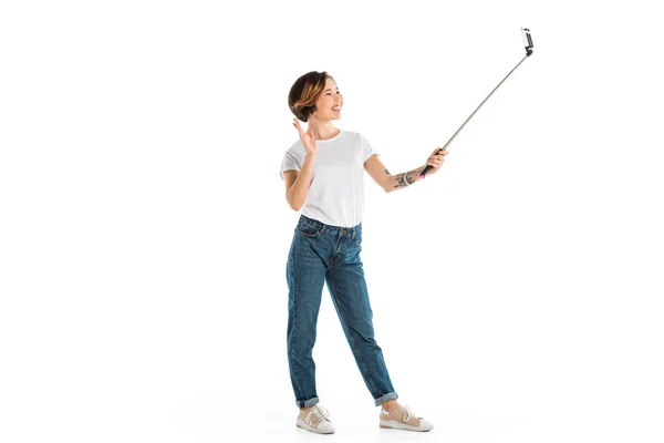 Souriant jeune femme agitant, prendre selfie sur smartphone et en utilisant selfie bâton isolé sur blanc — Photo de stock