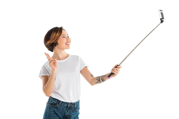 Atractiva mujer joven sonriente tomando selfie en el teléfono inteligente usando palo de selfie aislado en blanco - foto de stock