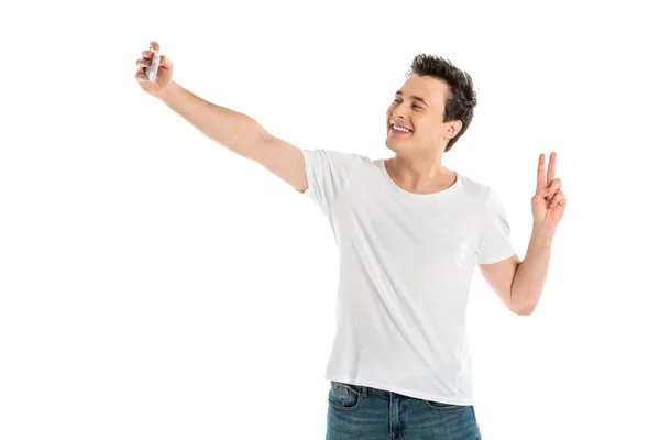 Bello uomo sorridente prendendo selfie su smartphone e mostrando segno di pace isolato su bianco — Foto stock