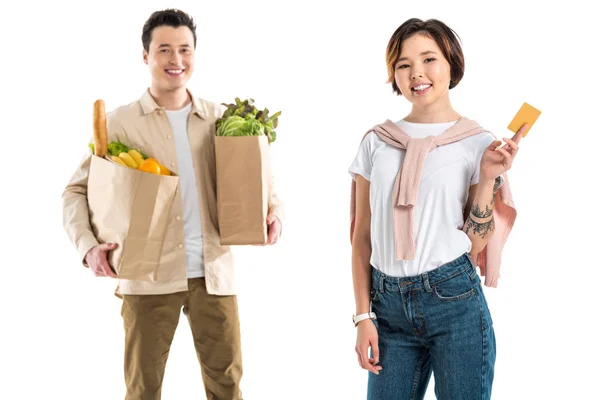 Sonriente marido sosteniendo bolsa de comestibles mientras esposa presentando tarjeta de crédito aislado en blanco - foto de stock