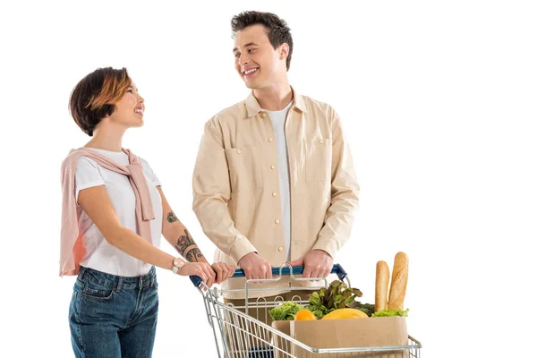 Lächelndes junges Paar mit Einkaufswagen voller Lebensmittel, die sich isoliert auf weißem Grund anschauen — Stockfoto