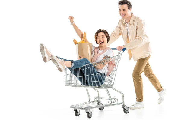 Smiling husband pushing shopping cart with wife inside holding groceries isolated on white, couple having fun — Stock Photo