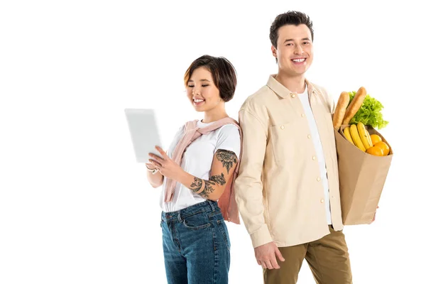 Cheerful husband holding grocery bag while wife using digital tablet isolated on white — Stock Photo