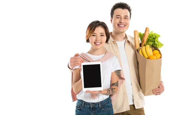 Улыбающийся муж держит пакет с продуктами, в то время как жена представляет цифровой планшет с чистым экраном изолированы на белом — стоковое фото