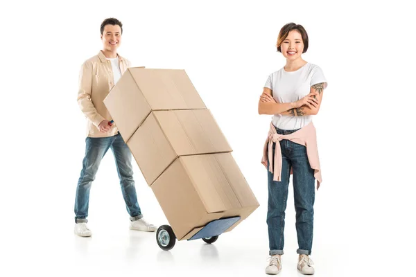 Marido llevando camión de mano con cajas de cartón en el fondo, mientras que la esposa mirando a la cámara, se mueve a nuevo concepto de casa - foto de stock