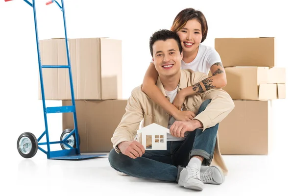 Пара обіймає і тримає модель будинку з картонними коробками на фоні, переходячи до нової концепції будинку — стокове фото