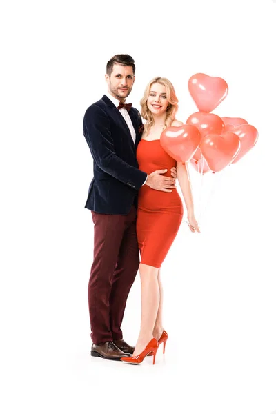 Hermosa pareja sonriente abrazándose con globos rojos mientras mira a la cámara aislada en blanco - foto de stock
