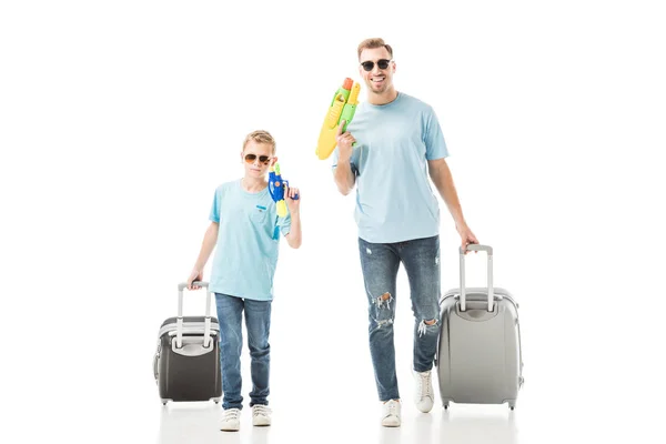 Padre e hijo caminando en gafas de sol y sosteniendo pistolas de agua y equipaje aislados en blanco - foto de stock