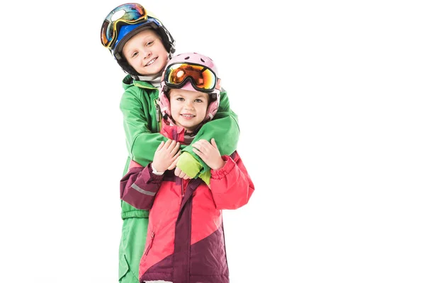 Hermana y hermano de pie y abrazándose en trajes de nieve aislados en blanco - foto de stock