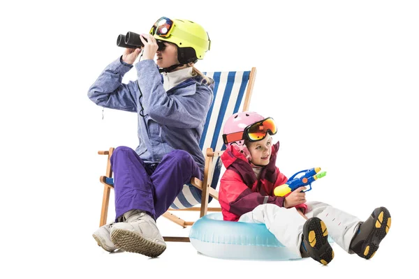 Frau im Skianzug mit Fernglas im Liegestuhl sitzend, während Tochter mit Wasserpistole auf Schwimmring sitzend isoliert auf weiß — Stockfoto