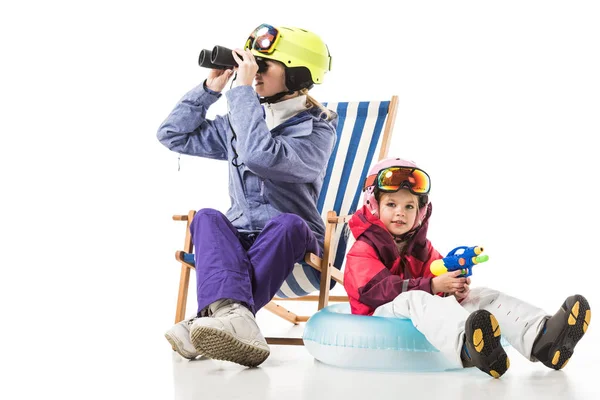 Woman with binoculars sitting in deck chair while daughter with water gun sitting on swim ring isolated on white — Stock Photo