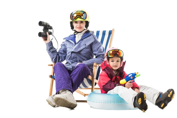 Mulher nova no terno de esqui com binóculos na cadeira de convés e filha pré-escolar com arma de água olhando para a câmera isolada no branco — Fotografia de Stock
