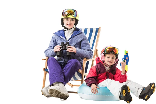 Donna sorridente con binocolo seduta sulla sedia a sdraio mentre figlia con pistola ad acqua che guarda la macchina fotografica isolata sul bianco — Foto stock