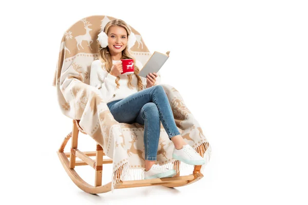 Молода усміхнена жінка тримає книгу і чашку кави, сидячи в кріслі-гойдалці ізольовано на білому — стокове фото