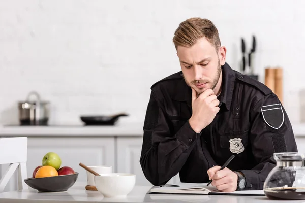 Thoughtful policeman writing in notebook at kitchen table — Stock Photo