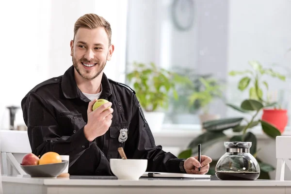 Policier souriant écrivant dans un carnet et mangeant des pommes à la table de la cuisine — Photo de stock