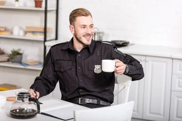 Schöner Polizeibeamter sitzt am Küchentisch, lächelt und trinkt Kaffee — Stockfoto