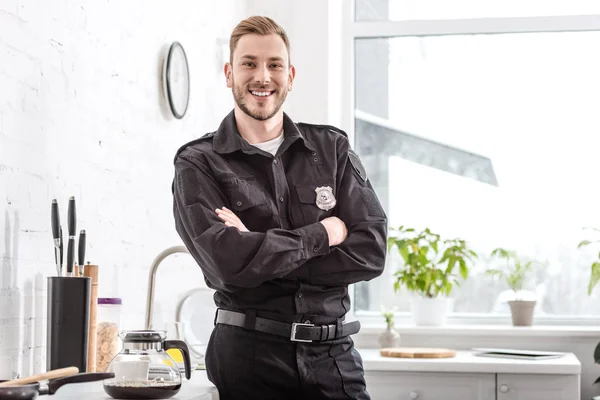 Sorridente policial com braços cruzados de pé ao lado da mesa da cozinha — Fotografia de Stock