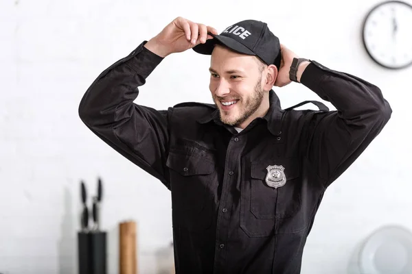 Apuesto oficial de policía sonriendo y poniéndose gorra en la cocina - foto de stock