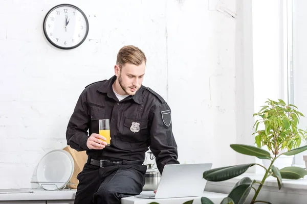 Jeune policier assis sur la table avec un verre de jus d'orange et utilisant un ordinateur portable à la cuisine — Photo de stock