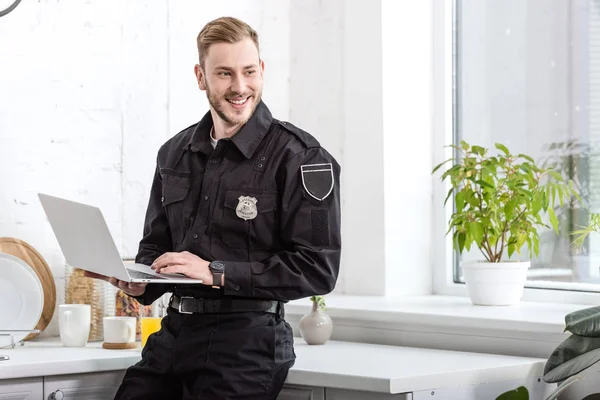 Beau policier souriant et utilisant un ordinateur portable à la cuisine — Photo de stock