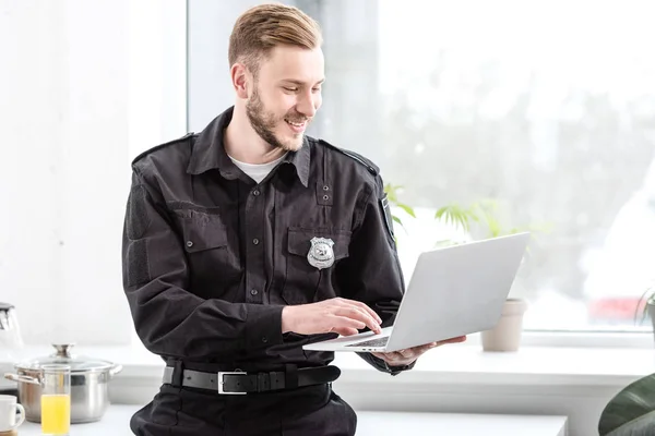 Улыбающийся офицер полиции, стоящий и пользующийся ноутбуком у окна кухни — стоковое фото