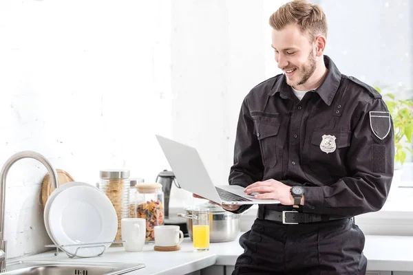 Policier souriant debout et utilisant un ordinateur portable à la cuisine — Photo de stock