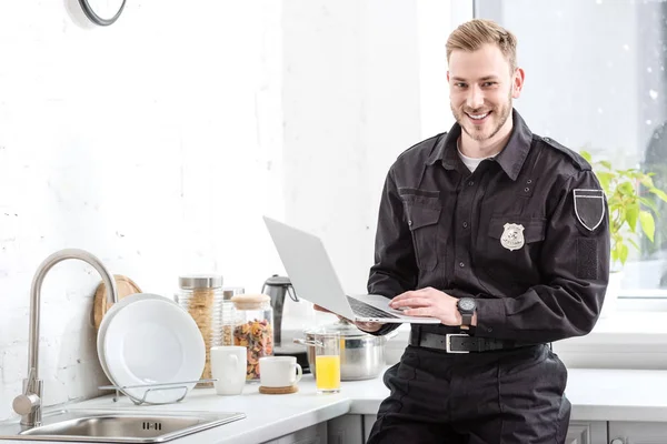 Улыбающийся полицейский стоит с ноутбуком на кухне — стоковое фото