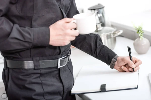 Vista cortada de policial segurando xícara de café e escrevendo em caderno na mesa da cozinha — Fotografia de Stock