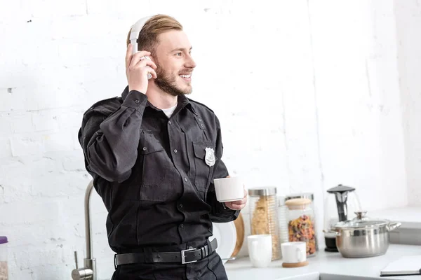 Schöner Polizist, der Kaffee trinkt und in der Küche mit Kopfhörern Musik hört — Stockfoto