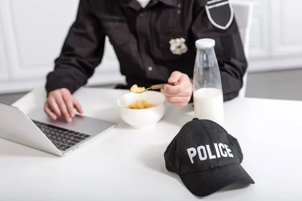 Vista recortada del oficial de policía utilizando el ordenador portátil y comer copos de maíz en la mesa de la cocina - foto de stock