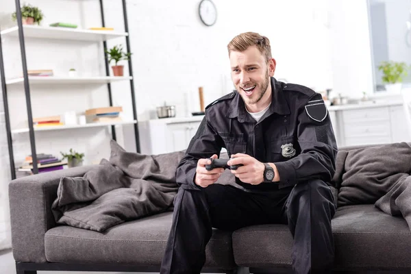 Rindo policial com gamepad sentado no sofá e jogando videogame — Fotografia de Stock