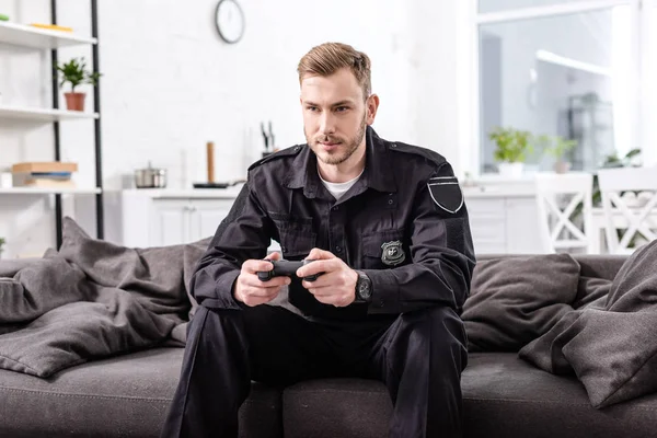 Концентрированный полицейский с геймпадом, сидящим на диване и играющим в видеоигры — стоковое фото