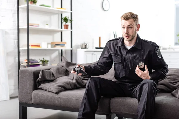 Surprised police officer sitting on couch with gamepad and playing video game — Stock Photo