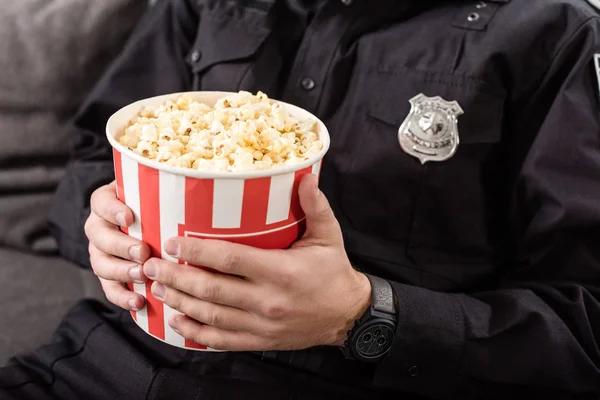 Обрезанный вид полицейского, сидящего на диване с полосатым ведром попкорна — стоковое фото