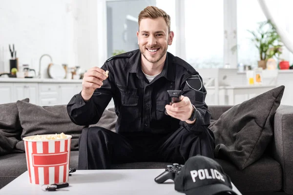 Офицер полиции сидит на диване, держит пульт дистанционного управления и ест попкорн — стоковое фото