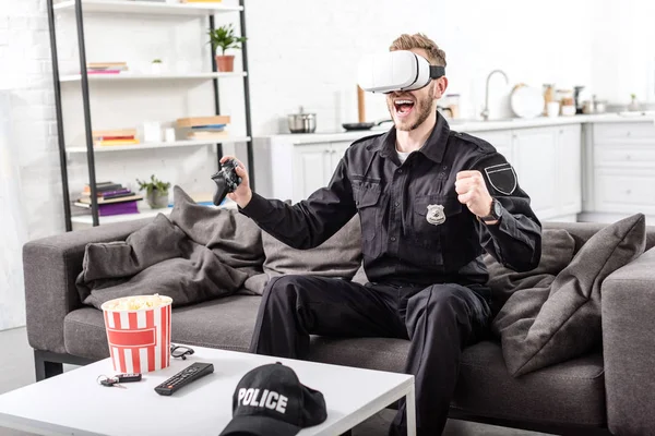 Полицейский с наушниками виртуальной реальности на голове, играющий в видеоигру и кричащий от радости на диване — стоковое фото