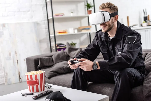 Policeman with virtual reality headset on head holding gamepad, sitting on couch and playing video game — Stock Photo