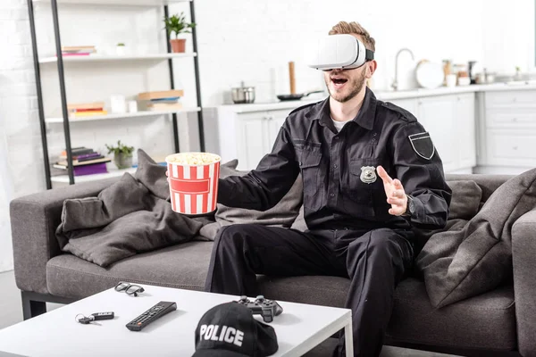 Полицейский с наушниками виртуальной реальности на голове, держащий ведро попкорна и сидящий на диване — стоковое фото