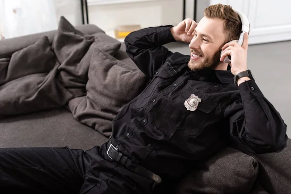 Улыбающийся полицейский сидит на диване и надевает наушники — стоковое фото