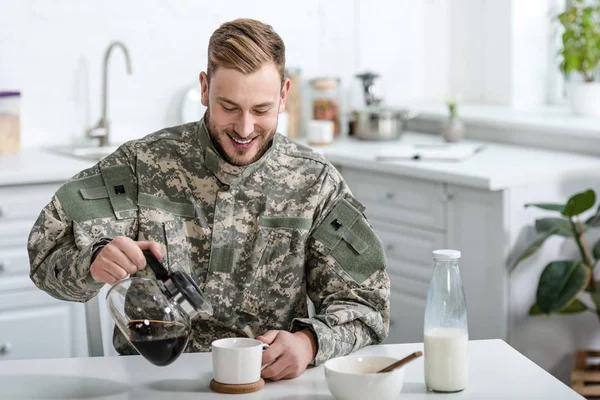 Schöner Soldat in Uniform lächelt und schenkt Kaffee in Tasse in Küche ein — Stockfoto