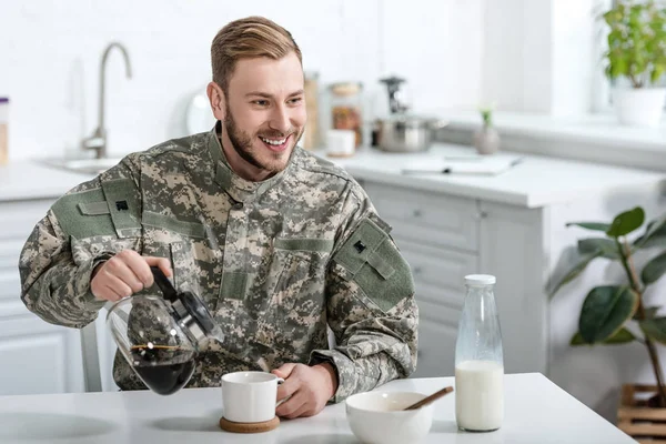 Beau homme en uniforme militaire souriant et versant du café dans une tasse de bouilloire à la table de cuisine — Photo de stock