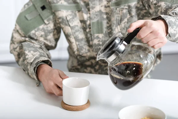 Abgeschnittene Ansicht eines Mannes in Tarnuniform, der Kaffee aus einem Wasserkocher in eine Tasse gießt — Stockfoto