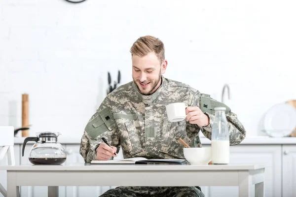 Uomo sorridente in uniforme mimetica che beve caffè e scrive nel taccuino in cucina — Foto stock