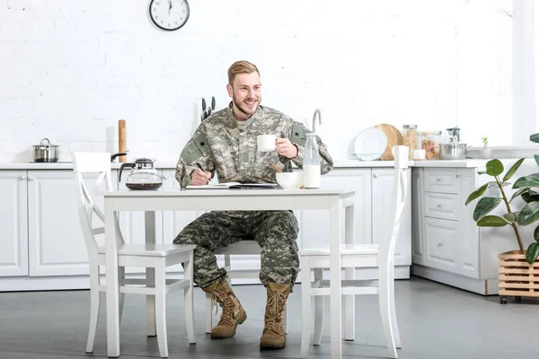 Handsome man in camouflage uniform writing in notebook and having breakfast at kitchen — Stock Photo