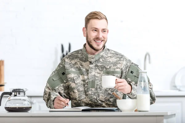 Hombre sonriente en uniforme de camuflaje escrito en cuaderno y desayunando - foto de stock