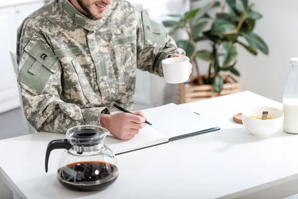 Vue recadrée de l'homme en uniforme militaire écrivant dans un cahier et buvant du café à table — Photo de stock